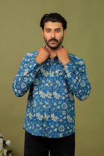 Load image into Gallery viewer, Man wearing a blue tropical floral printed full sleeve shirt with black jeans against an olive green background.