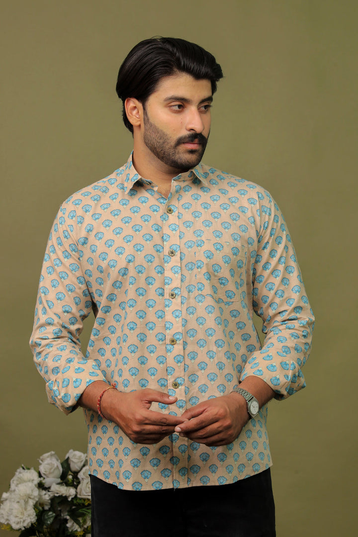 Smiling man wearing a beige cotton shirt with blue floral print and black jeans against an olive green background.