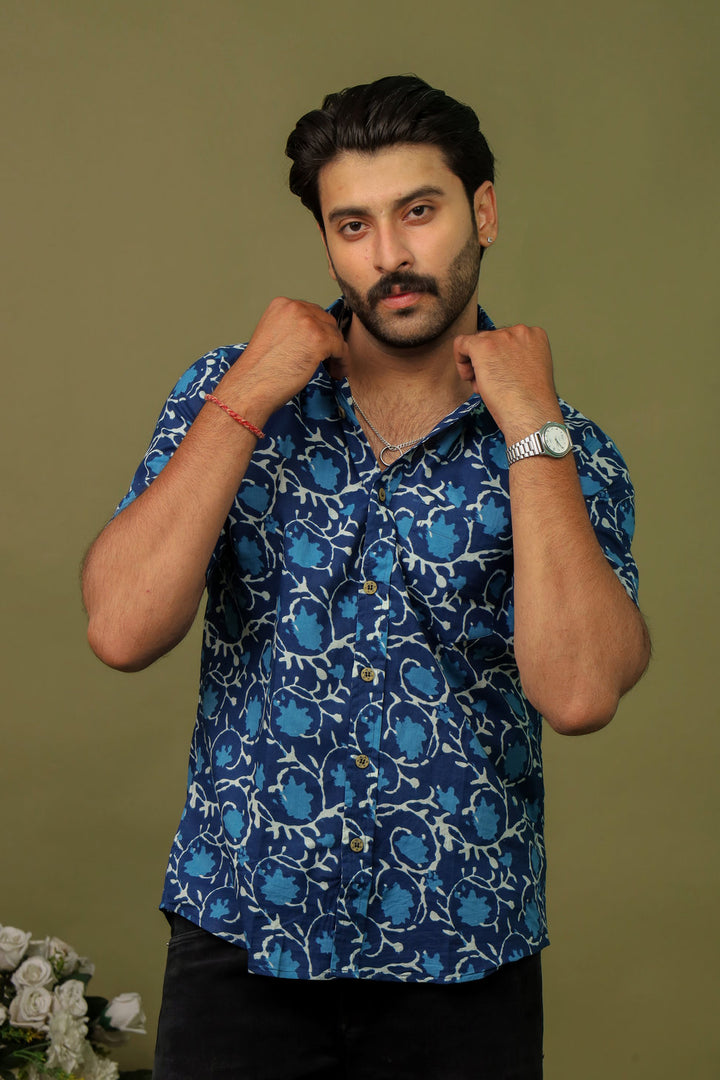 Man wearing a navy blue abstract printed half sleeve shirt with black jeans against an olive green background.