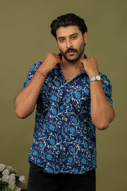 Man wearing a navy blue abstract printed half sleeve shirt with black jeans against an olive green background.
