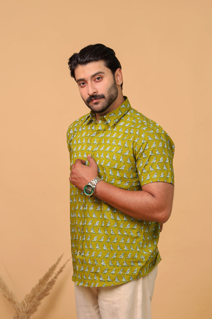 Man wearing an olive green bird printed half sleeve shirt with beige trousers against a beige background.
