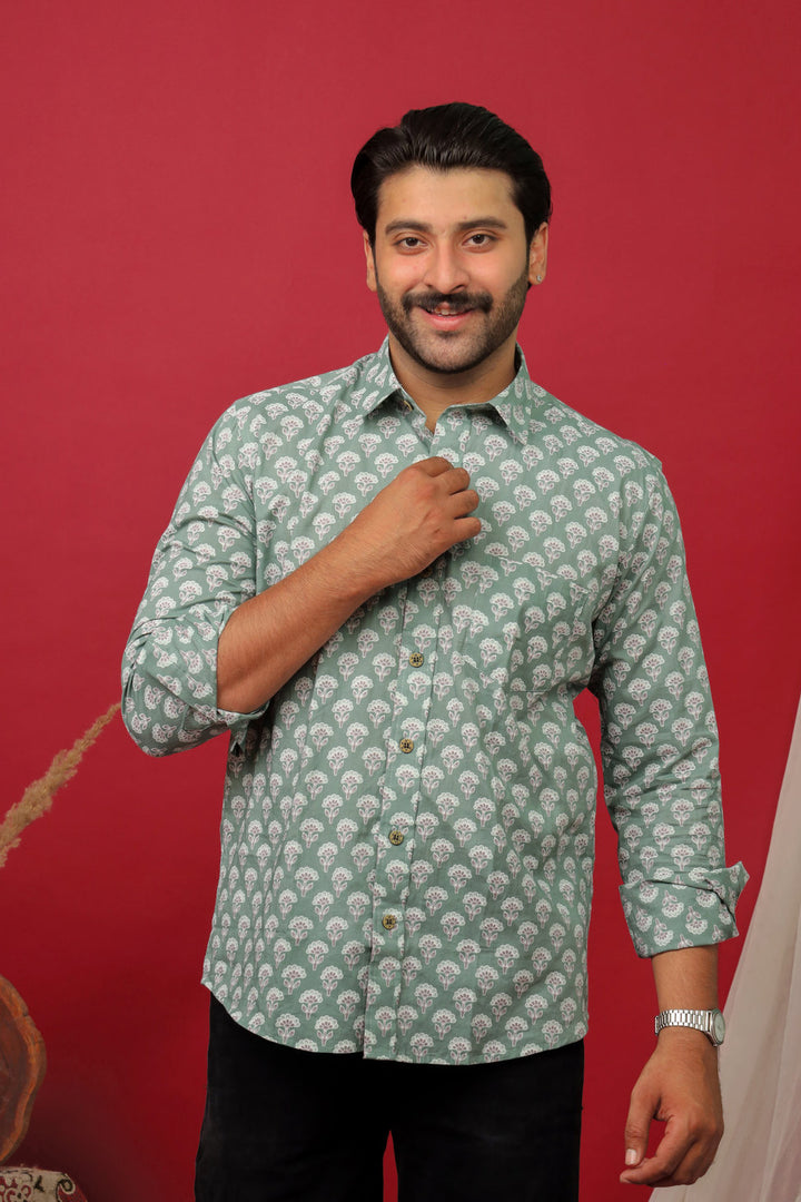 Man wearing a sage green floral printed full sleeve shirt with black jeans against a red background.