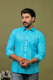 Man wearing a sky blue cotton shirt with subtle white Bandhani print and light denim jeans against an olive green background.