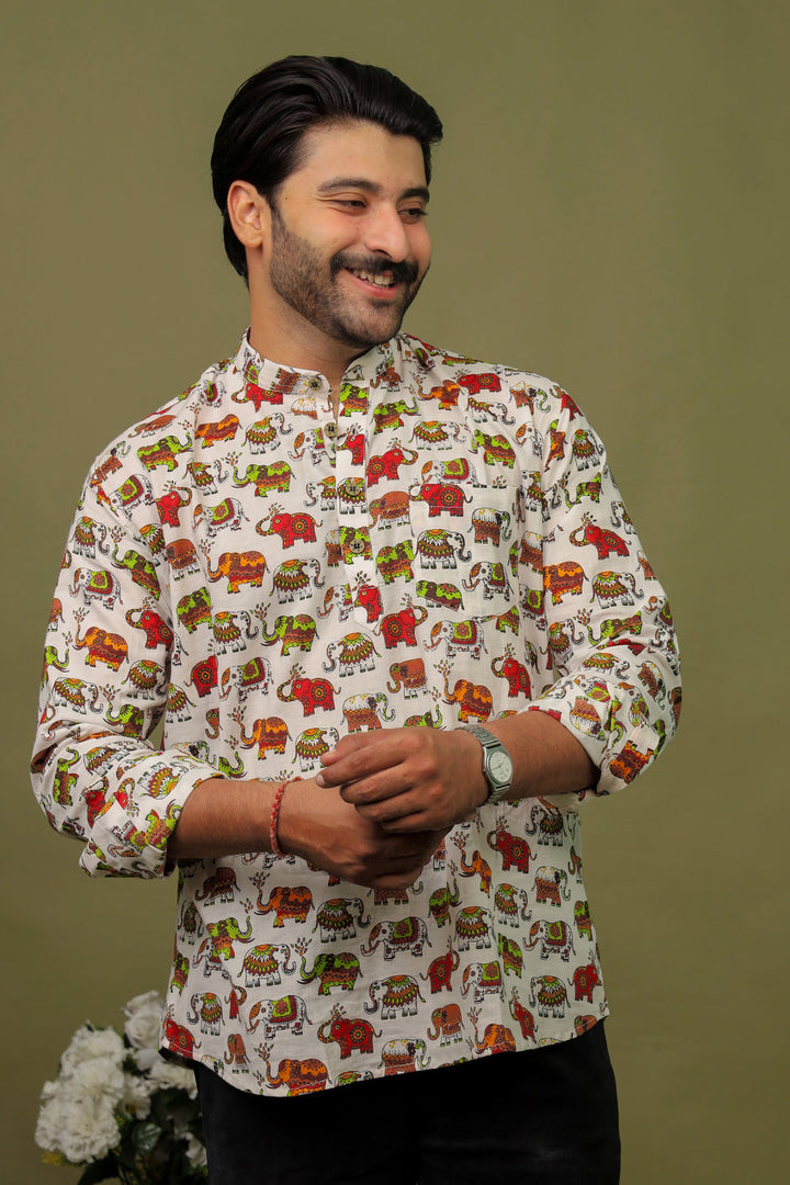 Smiling man wearing a white cotton shirt with colorful elephant print and black jeans against an olive green background.